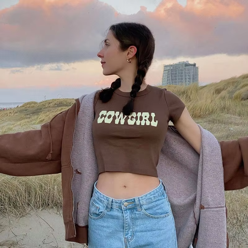 Brown cowgirl crop top with bold white lettering, short sleeves, and a trendy relaxed fit. Western-inspired casual fashion.